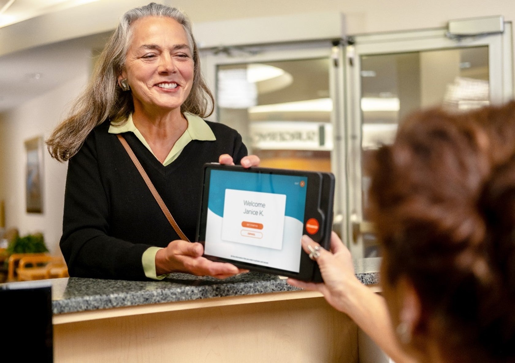 A photo of a patient smiling as she hands a Phreesia PadX to a front-desk healthcare worker.