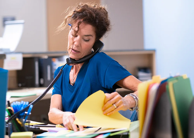 healthcare worker at front desk talking on phone while sifting through papers, appearing stressed and overwhelmed