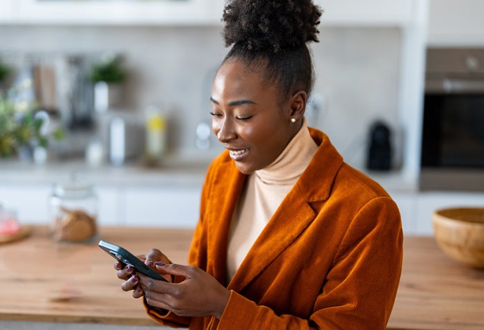 A woman wearing an orange jacket and holding a smartphone with both hands. She is looking at the screen and smiling.