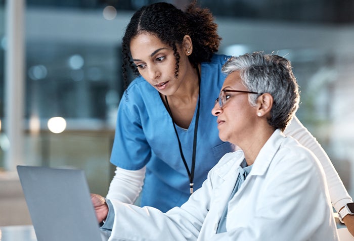 Two women looking at a laptop screen. One woman is wearing blue nurse scrubs, and the other is wearing a white-colored jacket. The woman wearing the jacket is pointing at something on the laptop screen.
