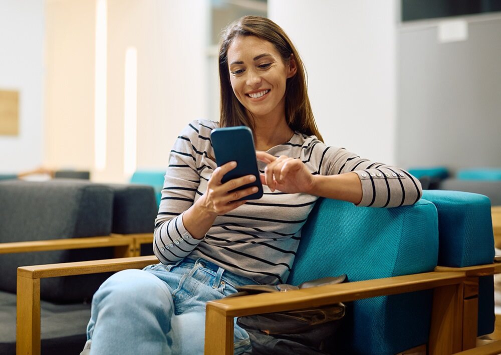 Woman sitting on a turquoise couch smiling while using a smartphone, wearing a striped shirt and jeans in a modern lounge area