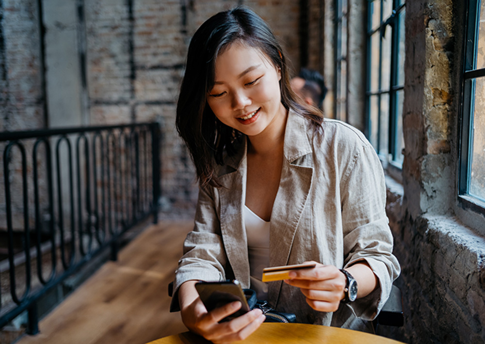 Woman smiling while holding a credit card and smartphone at an outdoor table in an urban alley, representing digital payment or healthcare transaction