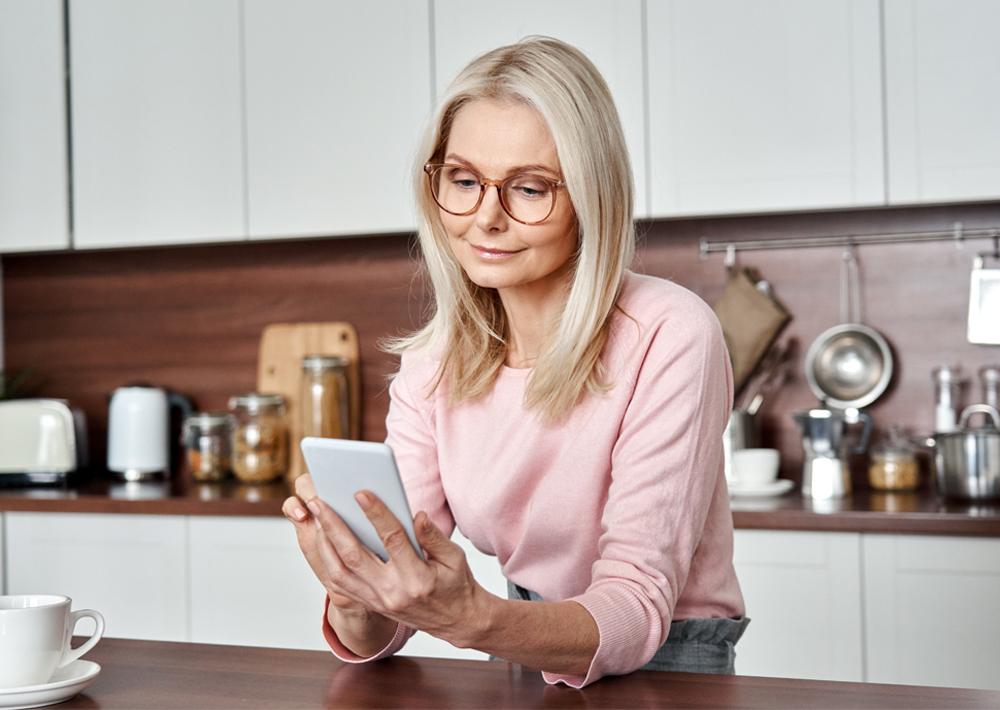 Woman wearing glasses and pink sweater looking at tablet in modern kitchen with white cabinets and dark backsplash