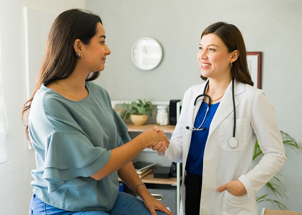 Female doctor in white coat shaking hands with female patient in medical office, both smiling during consultation