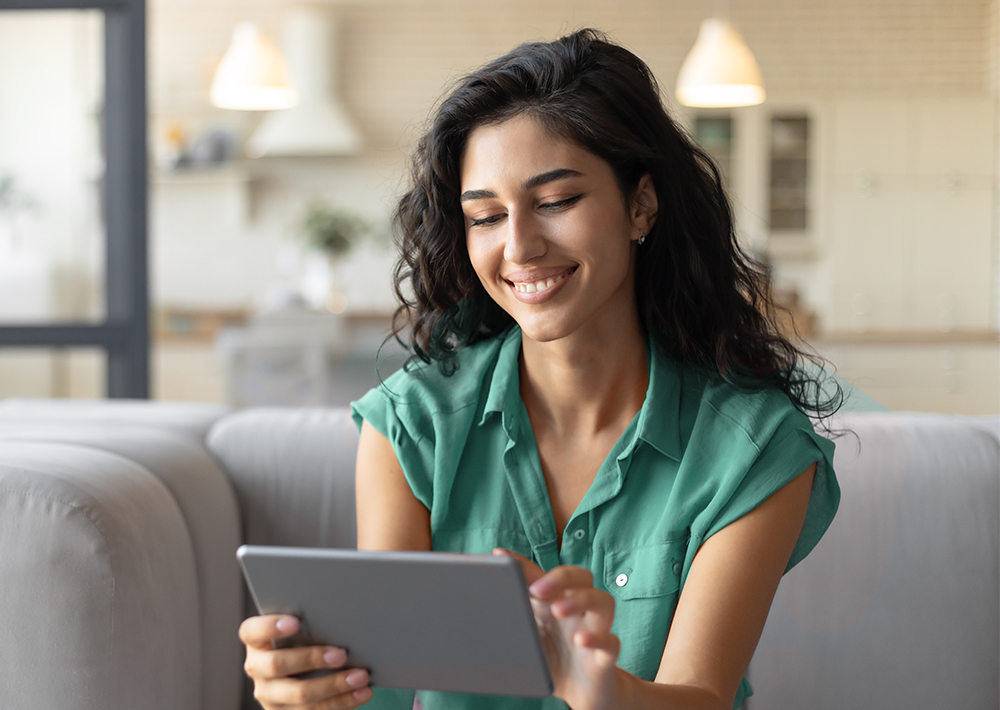 Smiling woman in green shirt sitting on gray couch, looking down at tablet device in her hands in bright, modern living room