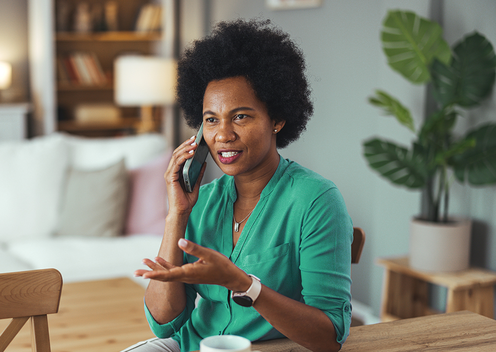 Woman in green shirt smiling while talking on phone at wooden desk in bright home office with plants and bookshelf in background