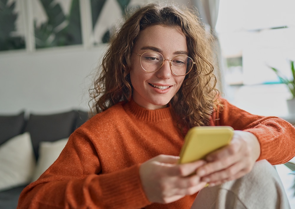 Smiling woman with curly brown hair and glasses wearing an orange sweater, holding a yellow phone while sitting on a couch in a bright office space