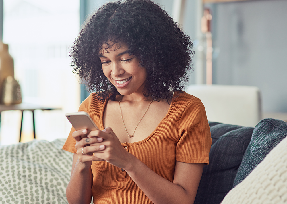 Smiling woman with curly dark hair wearing an orange ribbed top, looking at a mobile device while sitting on a couch in a bright, modern living room
