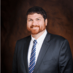 Professional headshot of a man with reddish-brown beard wearing a dark gray suit, white shirt, and navy striped tie against a brown background
