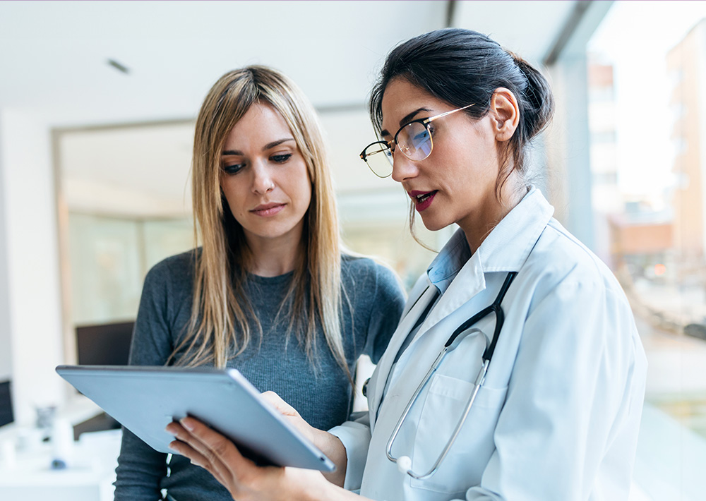 Female doctor in white coat and glasses reviewing tablet with patient in medical office setting