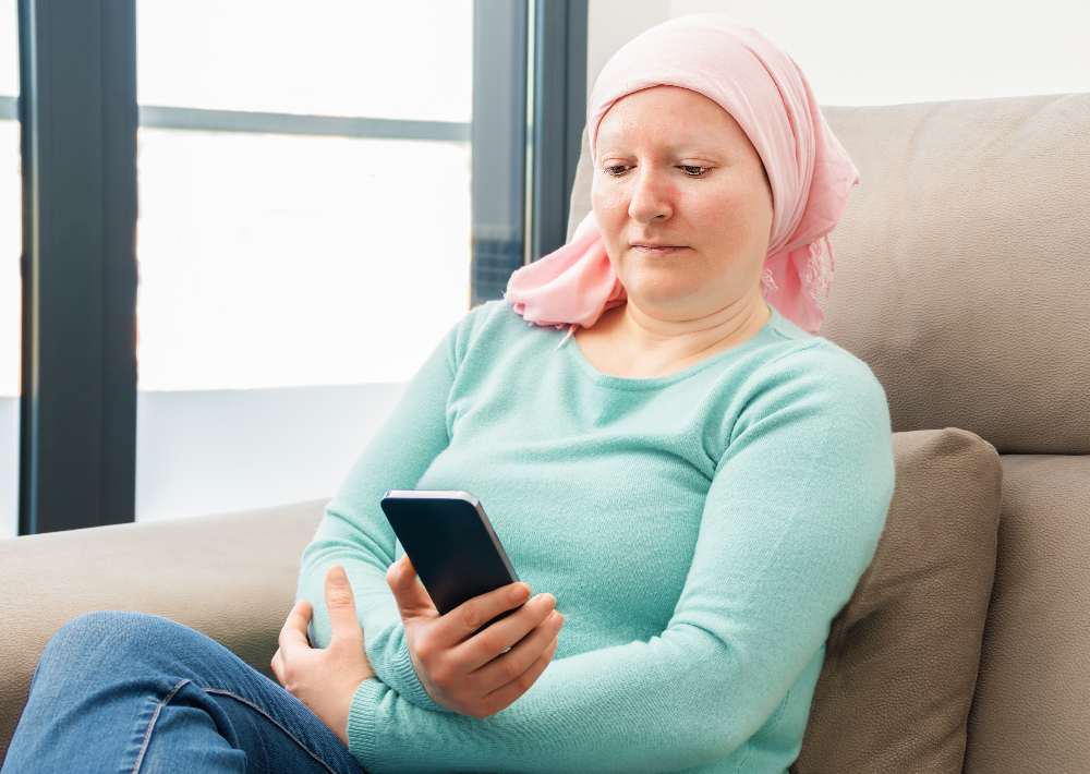 Woman wearing pink headscarf and teal sweater sitting on couch, looking at smartphone, with bright window in background