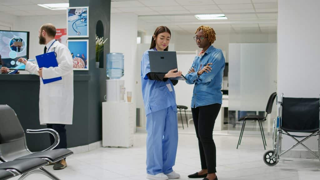 Healthcare worker in blue scrubs and visitor in denim shirt review laptop together in modern clinic reception area, with medical staff and water cooler visible in background