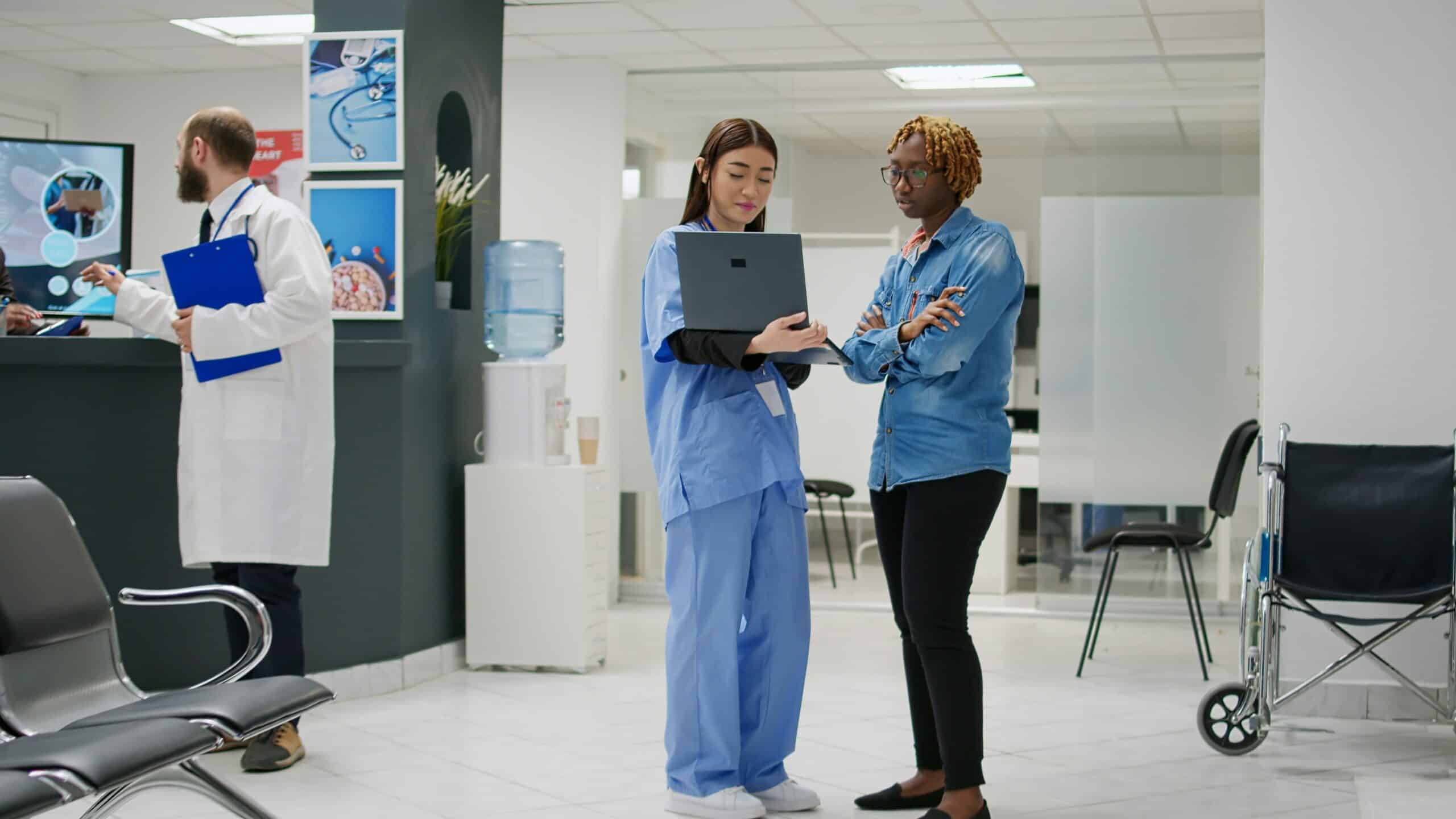 Healthcare worker in blue scrubs and visitor in denim shirt review laptop together in modern clinic reception area, with medical staff and water cooler visible in background