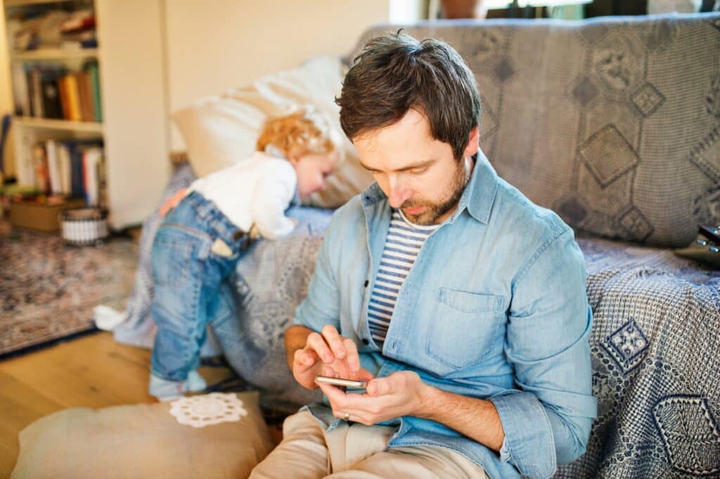 Man in denim shirt looking at smartphone while toddler plays on couch behind him in living room with bookshelves