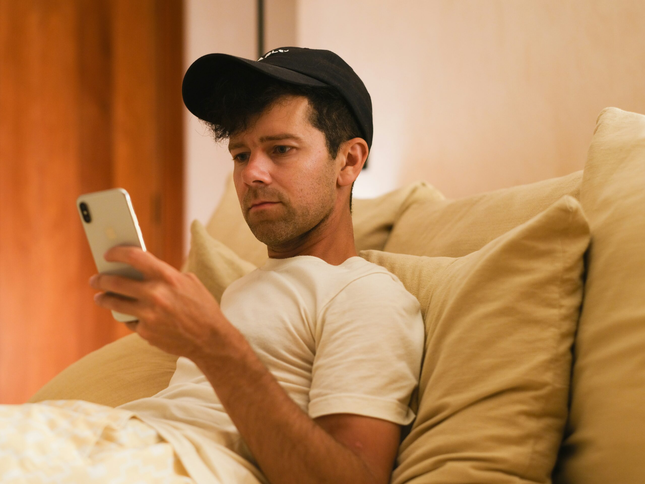 Man sitting on a beige couch wearing a black cap and white t-shirt, looking at his smartphone with a concerned expression