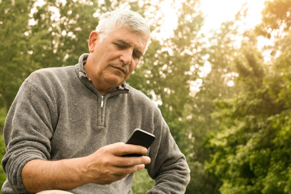Mature man in gray zip-up sweater looking down at smartphone outdoors among trees on a sunny day
