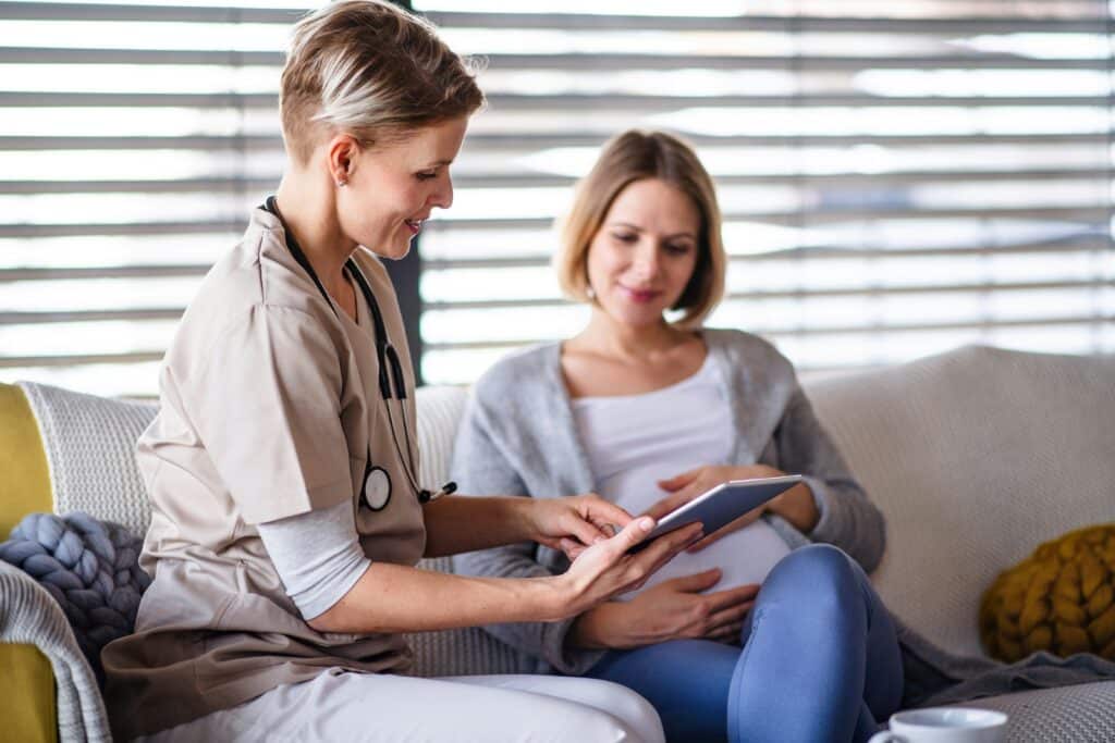 Healthcare provider in beige scrubs with stethoscope shows tablet to patient sitting on couch during medical consultation