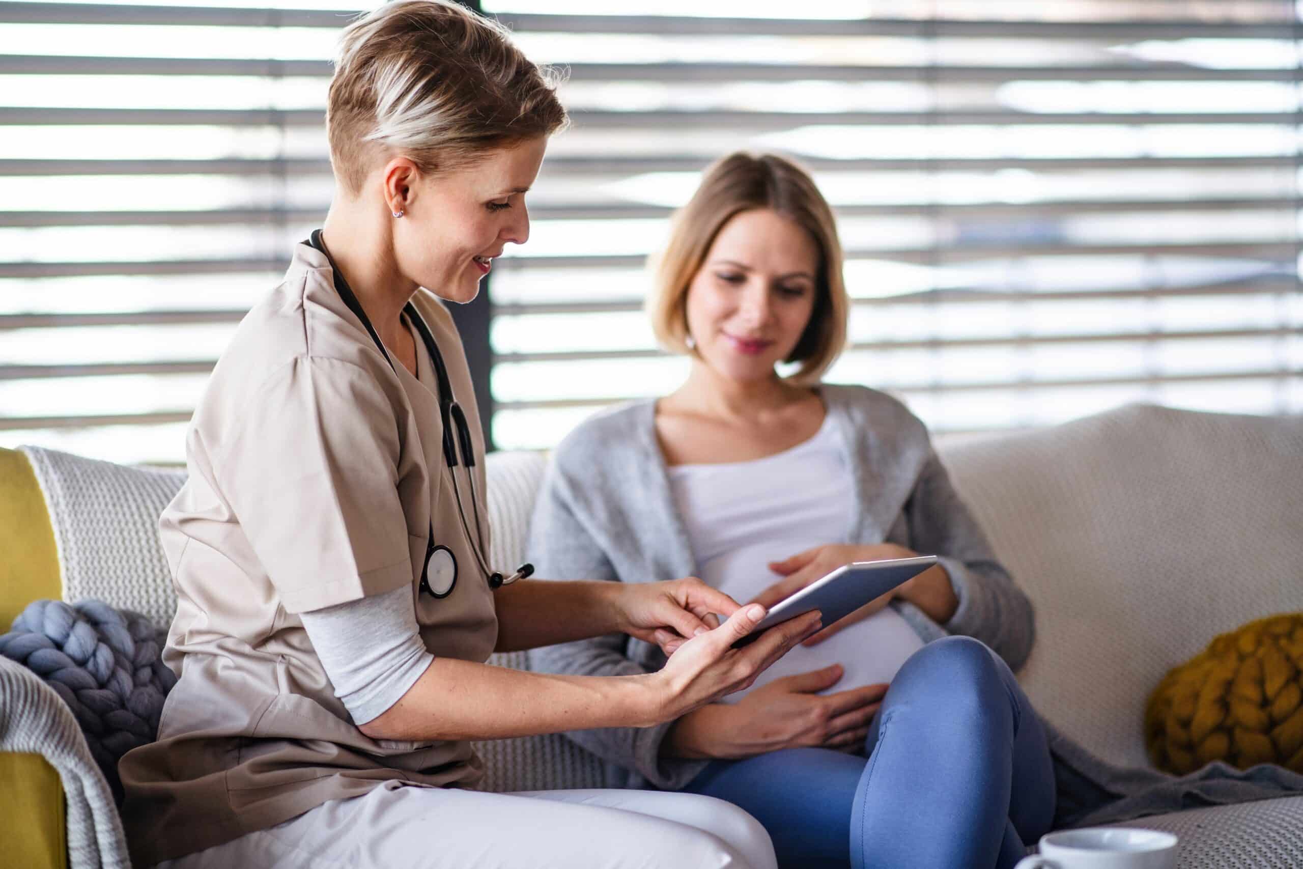 Healthcare provider in beige scrubs with stethoscope shows tablet to patient sitting on couch during medical consultation