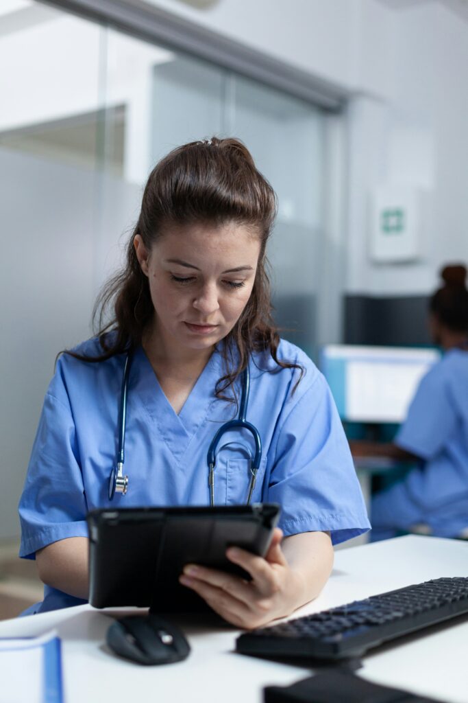 Healthcare nurse in blue scrubs using a tablet at desk with computer keyboard, stethoscope around neck, in modern clinical workspace
