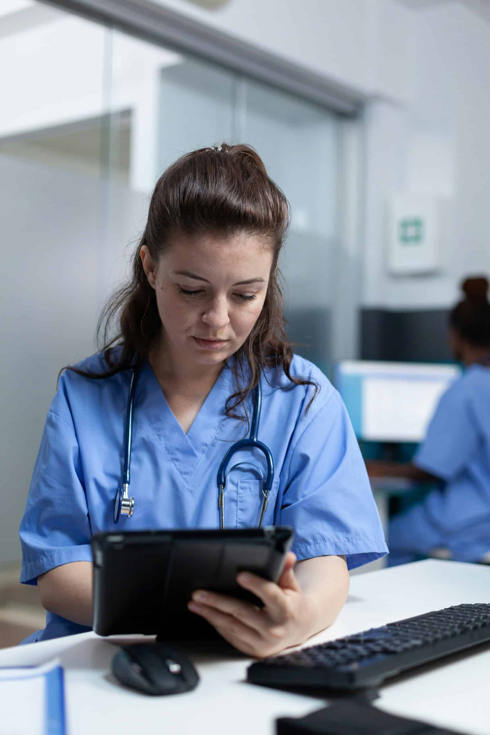 Healthcare nurse in blue scrubs using a tablet at desk with computer keyboard, stethoscope around neck, in modern clinical workspace