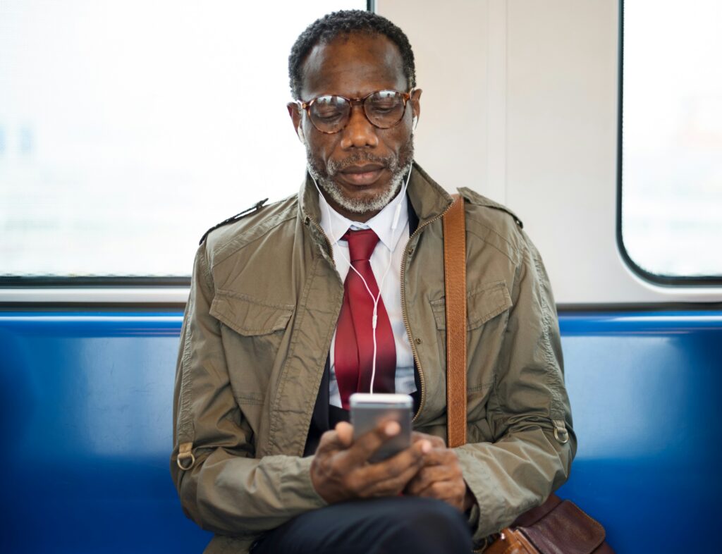 Older man wearing glasses and a tan jacket with red tie sitting on public transit while looking at his phone