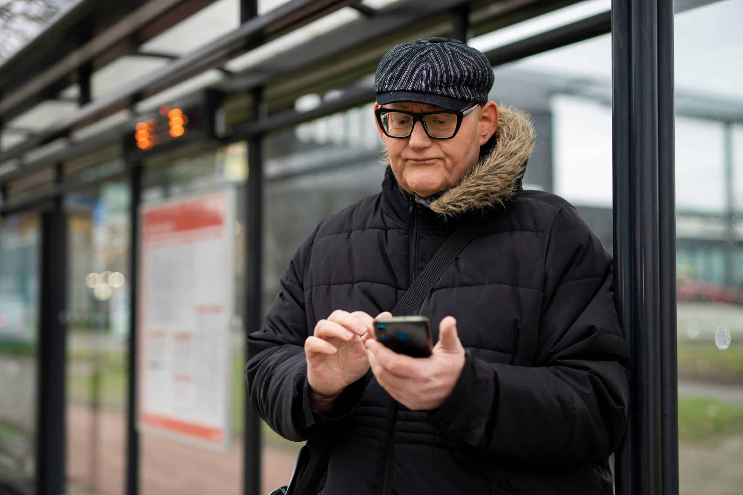 Older man wearing glasses and a black winter coat with fur-lined hood checks his phone while standing at a bus stop shelter