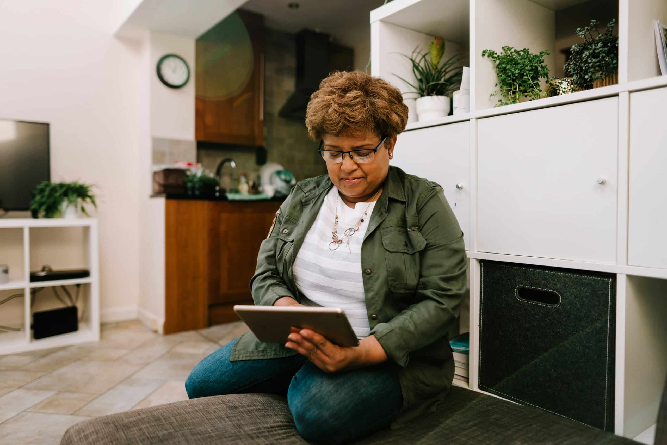 Woman in olive jacket sitting on floor cushion using tablet in modern home office with white storage shelving and potted plants