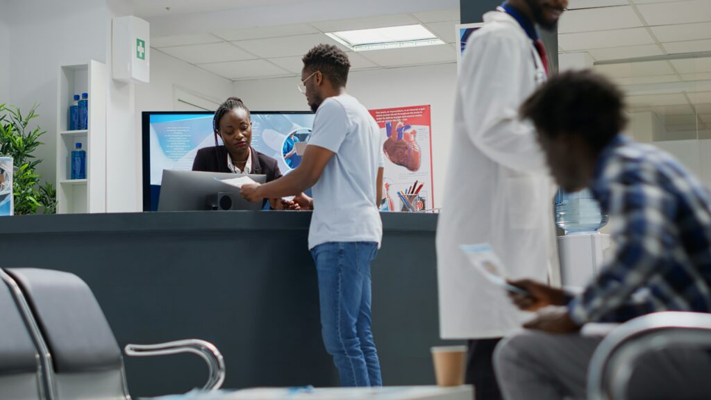 Patients checking in at a medical clinic reception desk with staff member at computer, anatomical posters visible on wall behind counter