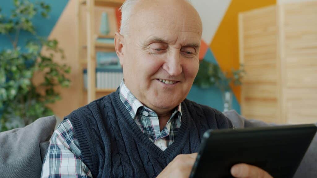 Senior man smiling while looking at a tablet device in a bright, modern room with colorful walls and indoor plants
