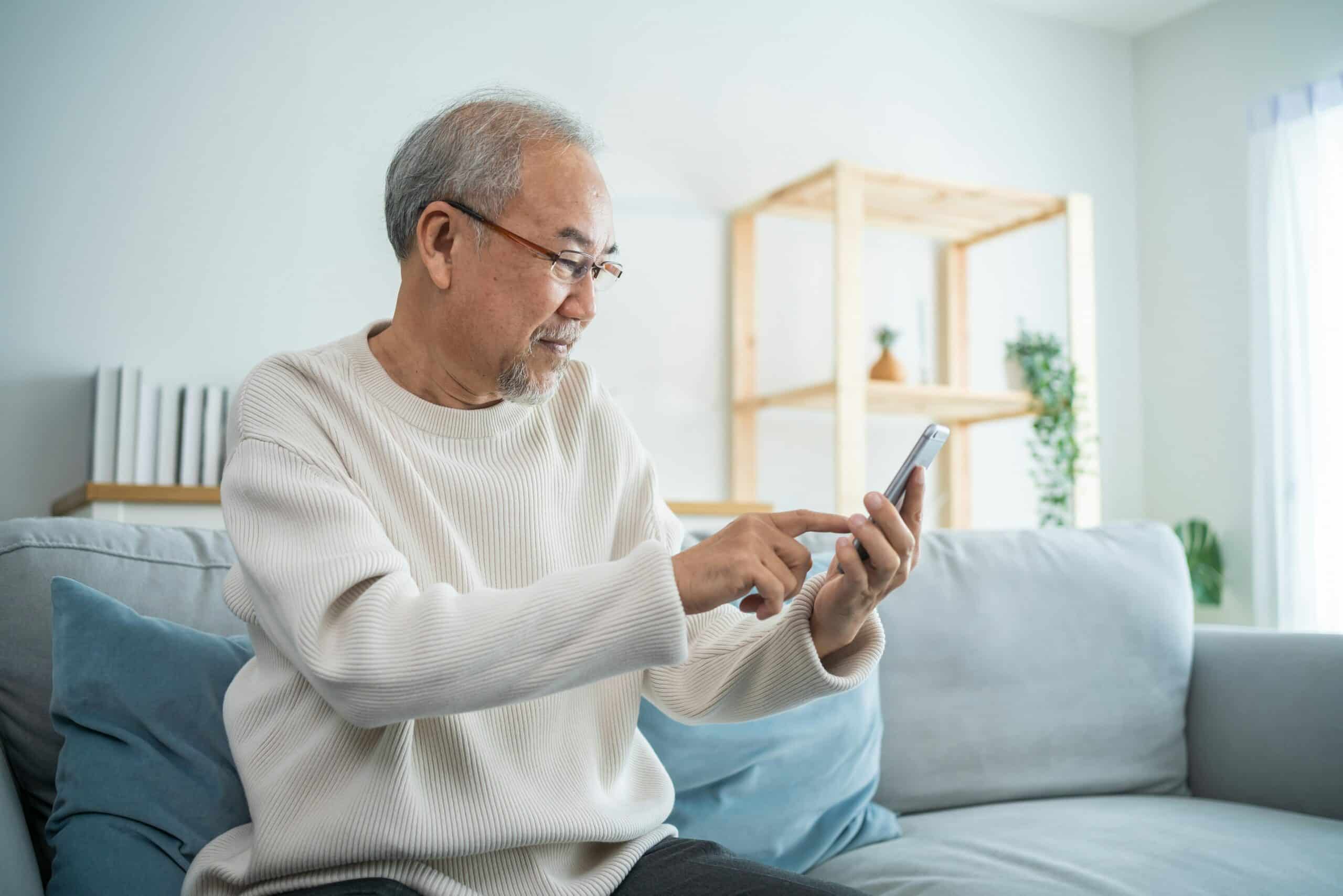 Senior man with glasses sitting on a gray couch, looking at a tablet device in his hands in a bright, modern living room