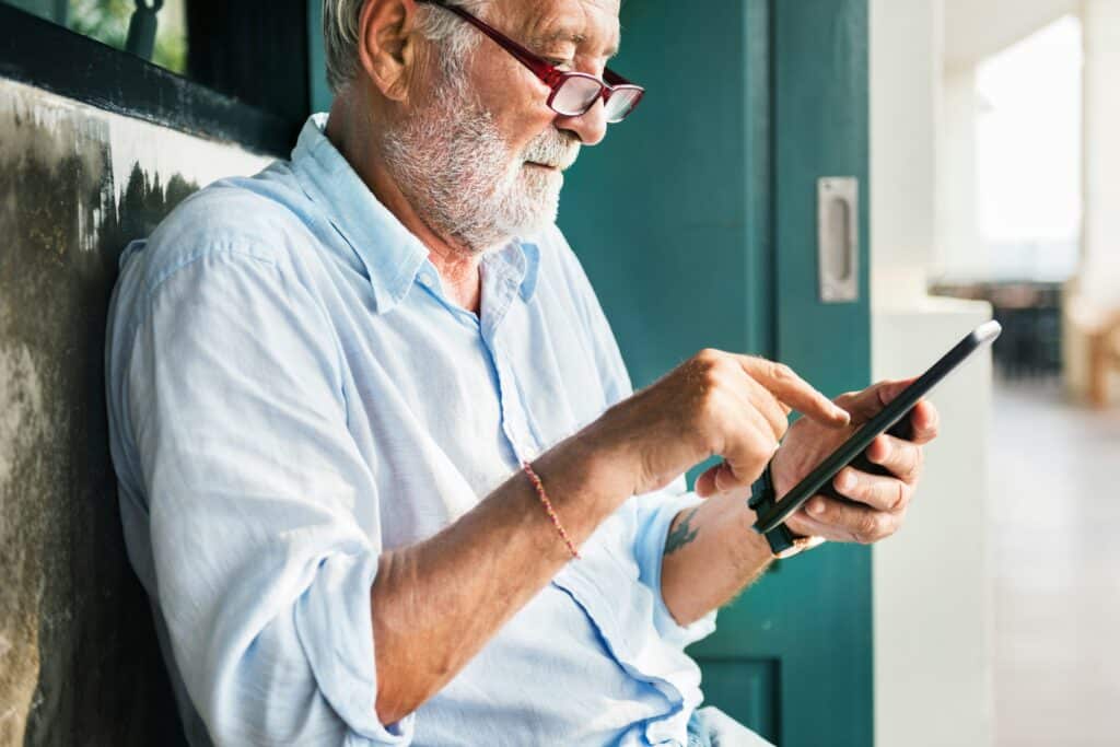 Older man with gray beard and glasses wearing light blue shirt, looking at smartphone while standing in doorway with teal-colored wall behind him