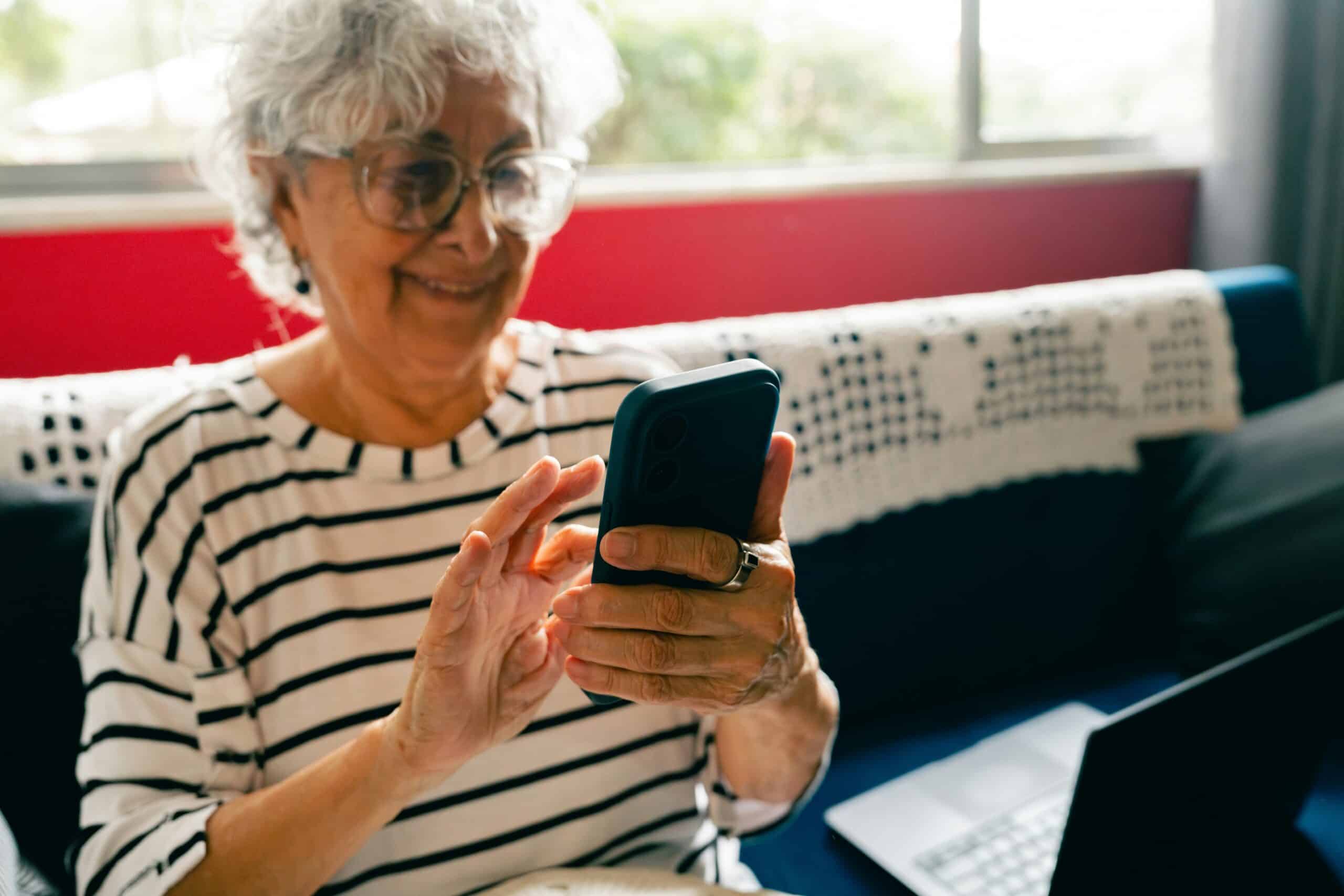 Smiling senior woman wearing glasses and striped shirt holds smartphone while sitting on red couch with laptop nearby