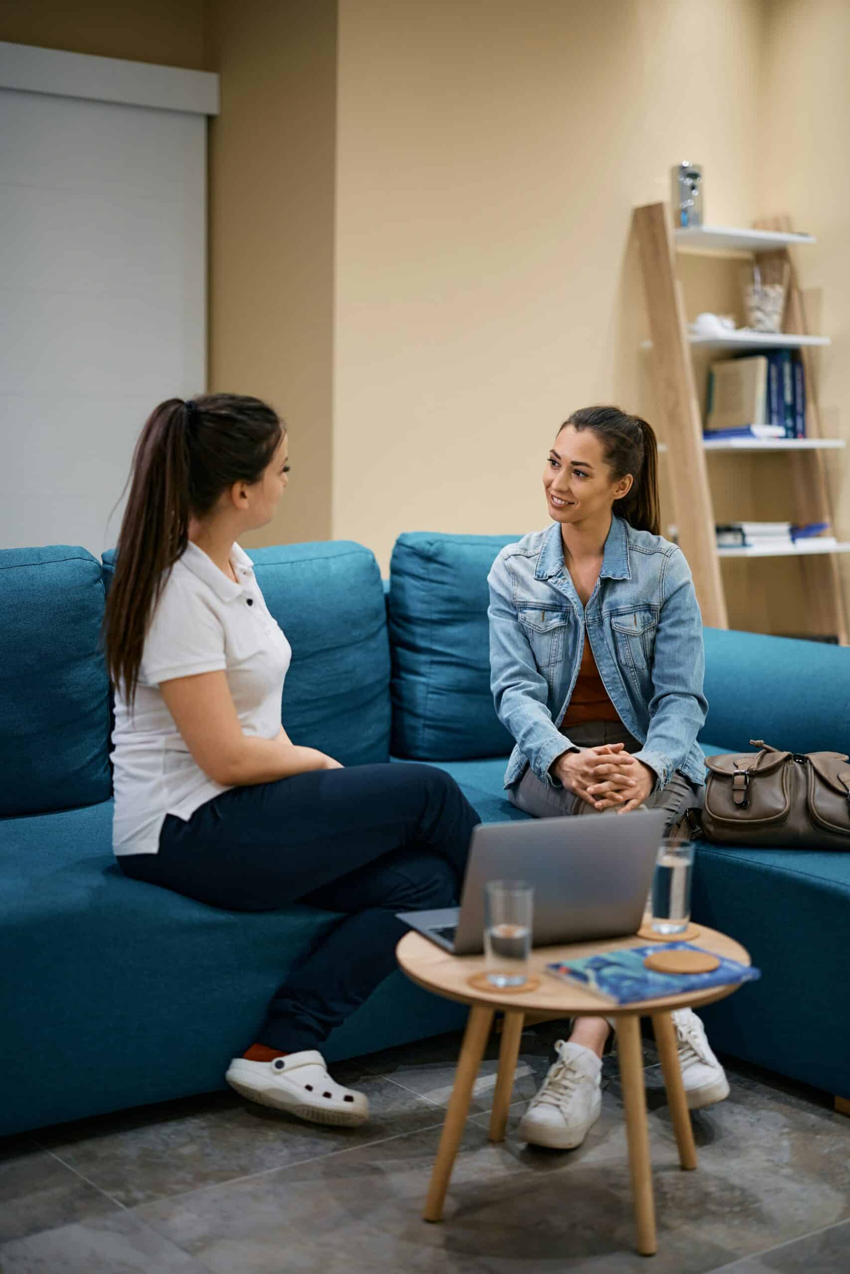 Two women sitting on a teal couch having a conversation in a modern waiting room, with a laptop and water glasses on a wooden table in front of them