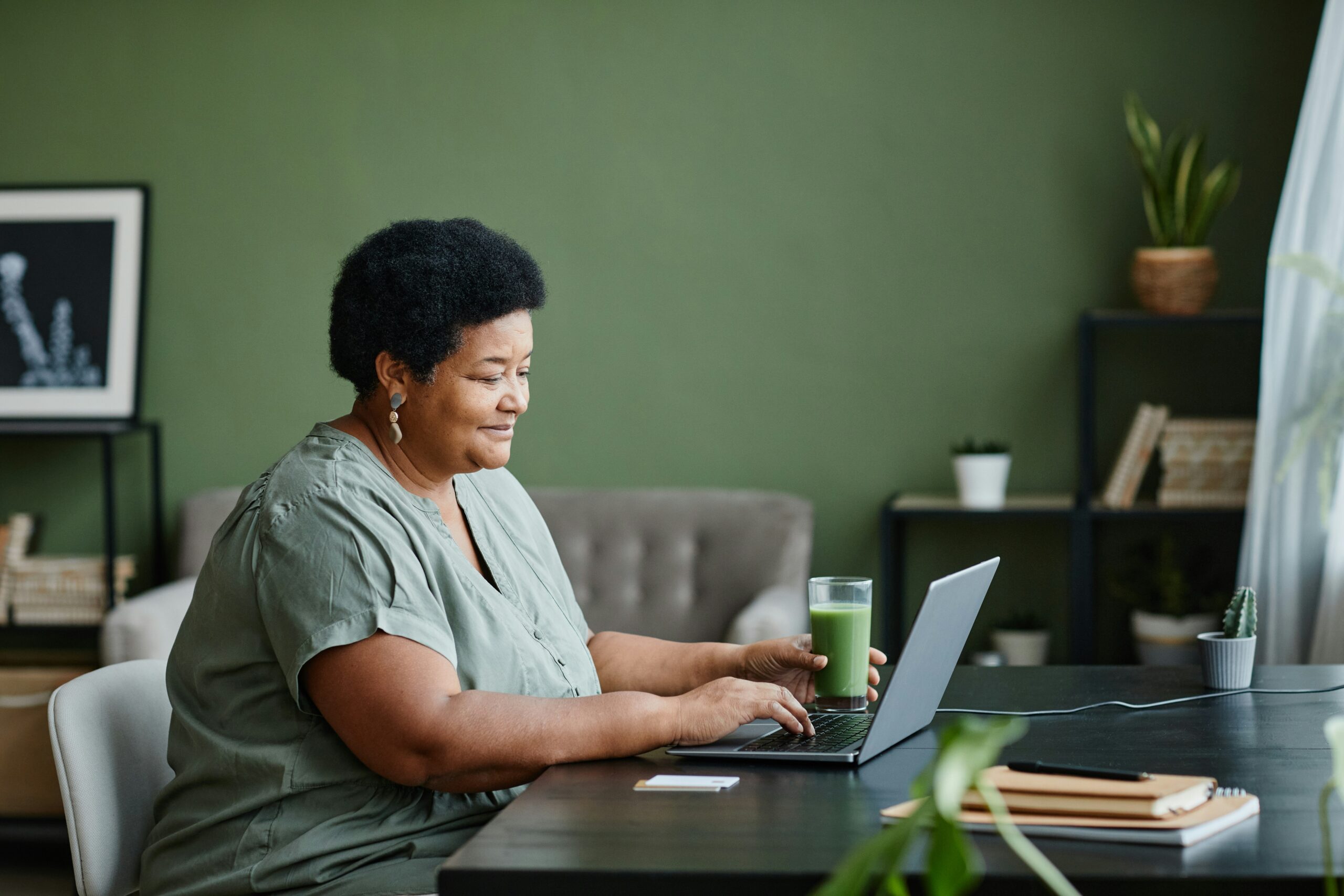Woman in sage green shirt working on laptop at desk in modern room with green walls, holding a green beverage, with potted plant and shelving in background
