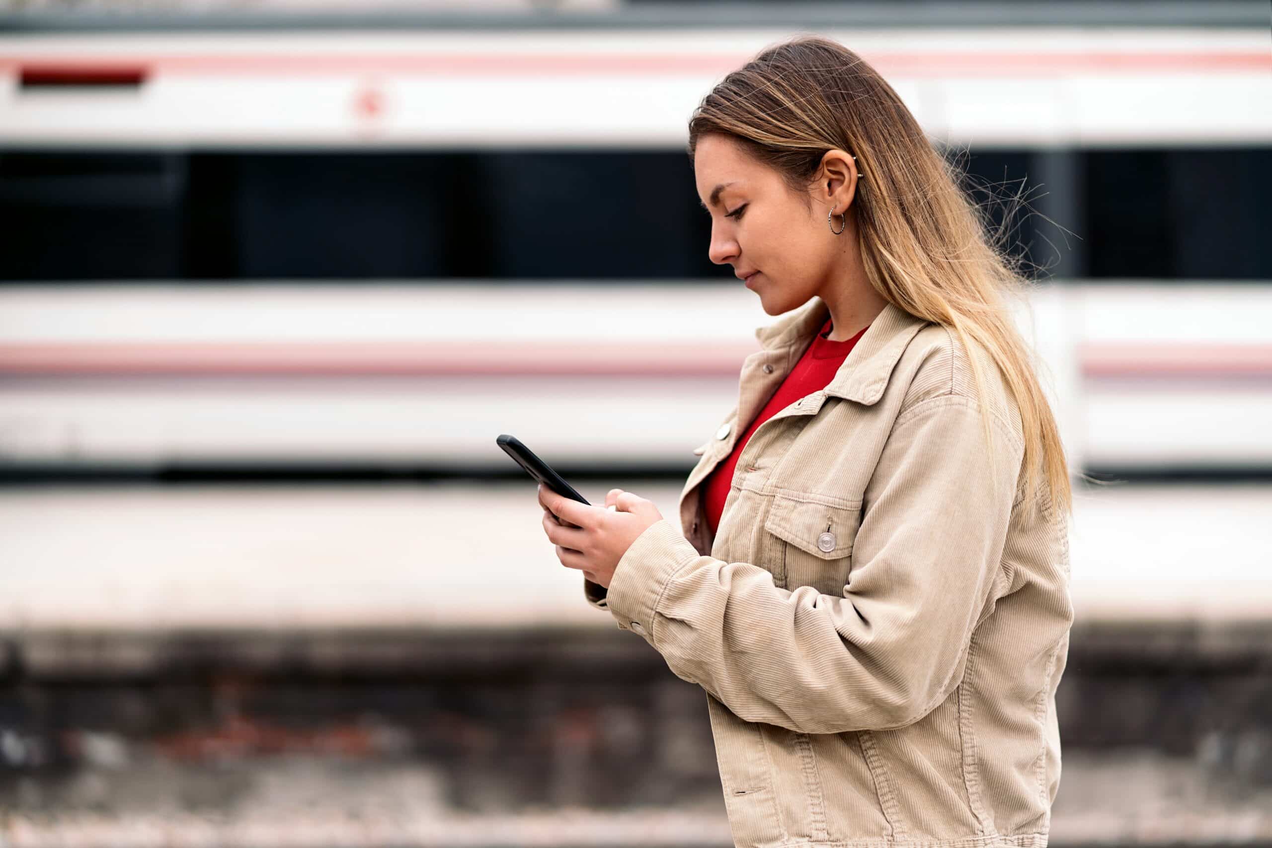 Woman in beige jacket and red shirt looking at smartphone while standing at a train platform with blurred trains in the background