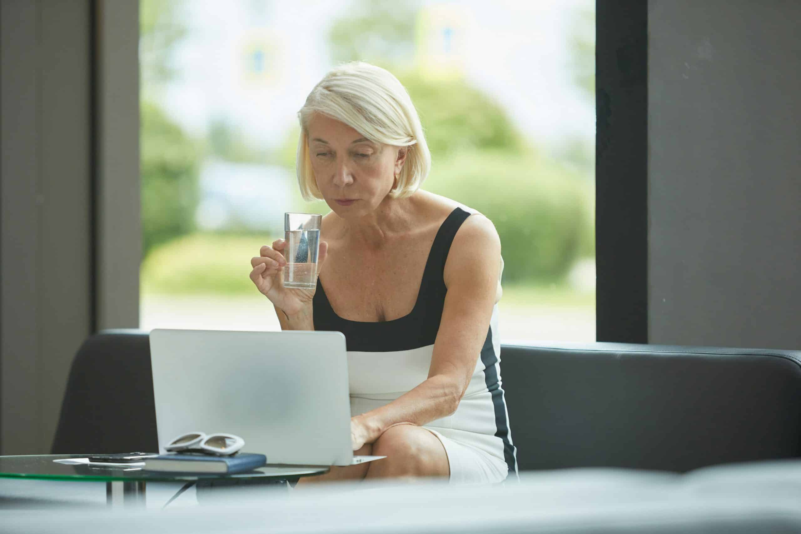 Woman with short blonde hair sitting at a desk with a laptop, holding a glass of water and looking at the screen. Sunlit windows and greenery visible in the background