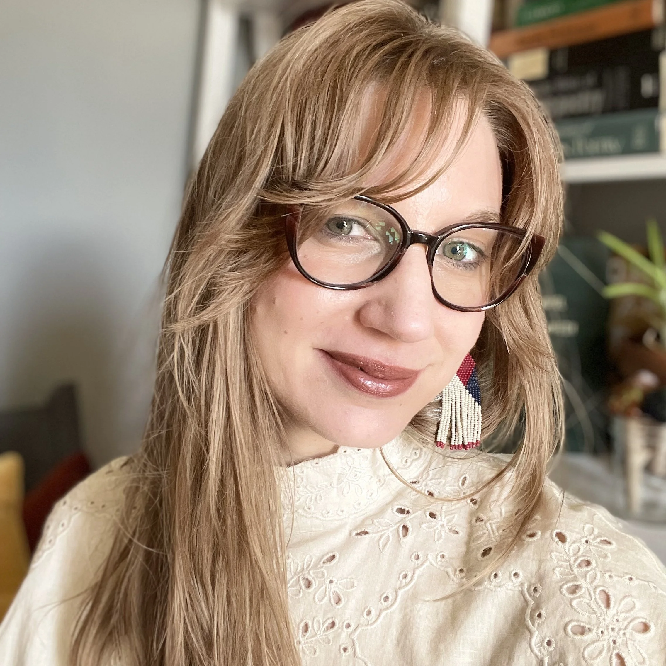 Woman with shoulder-length brown hair and bangs wearing black-framed glasses, a cream eyelet blouse, and colorful beaded earrings, smiling at the camera in an indoor setting