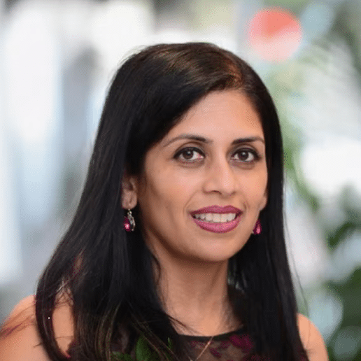 Professional headshot of Snedha Patel, a woman with long black hair, pink lipstick, and pink earrings, smiling at the camera against a blurred background