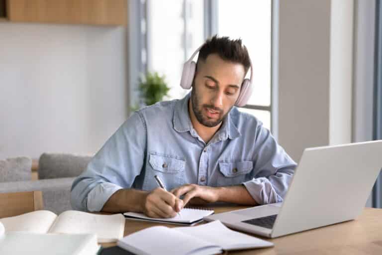 man with headphones on at desk writing notes in front of laptop