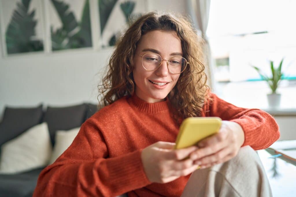 Smiling woman with curly brown hair and glasses wearing an orange sweater, holding a yellow phone while sitting on a couch in a bright office space